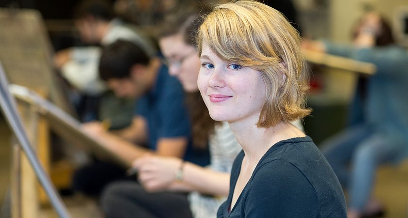 A young female student of Fine Arts smiles for the camera, her class can be seen at work in the background.